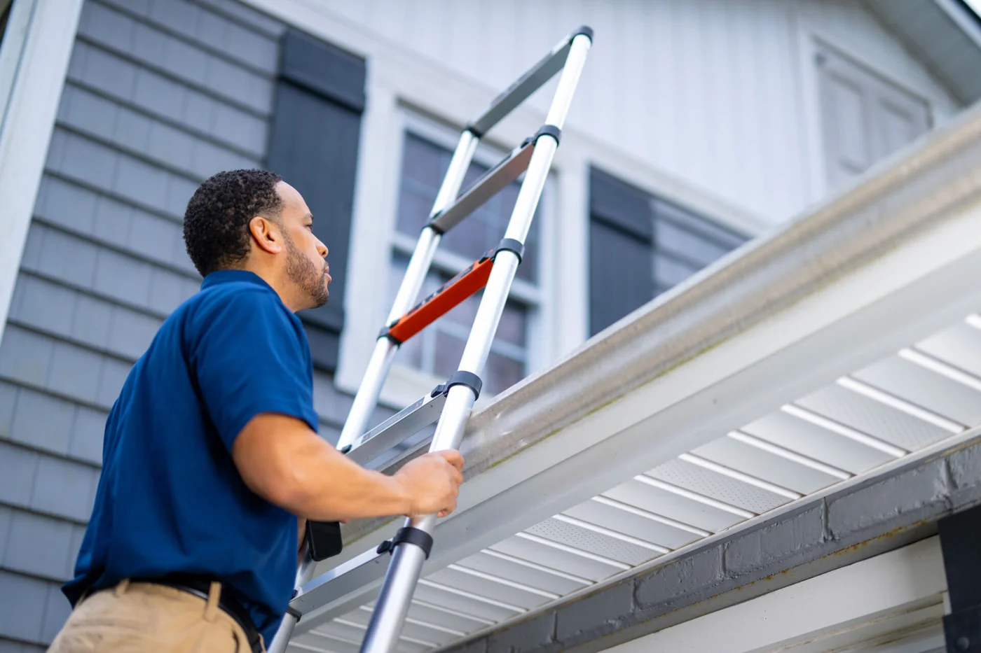 Home Genius Exteriors home remodeling expert climbing a ladder to perform a free roof inspection on a gray-sided house