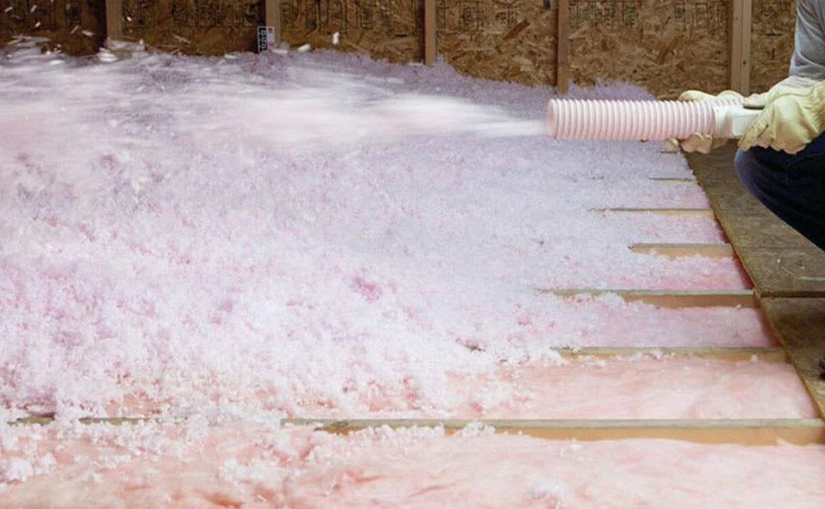 A technician blows loose pink fiberglass insulation across the attic floor joists through a hose.