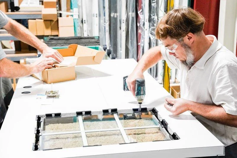 A craftsman assembles a ProVia door panel with an insulated glass insert at a factory workbench.
