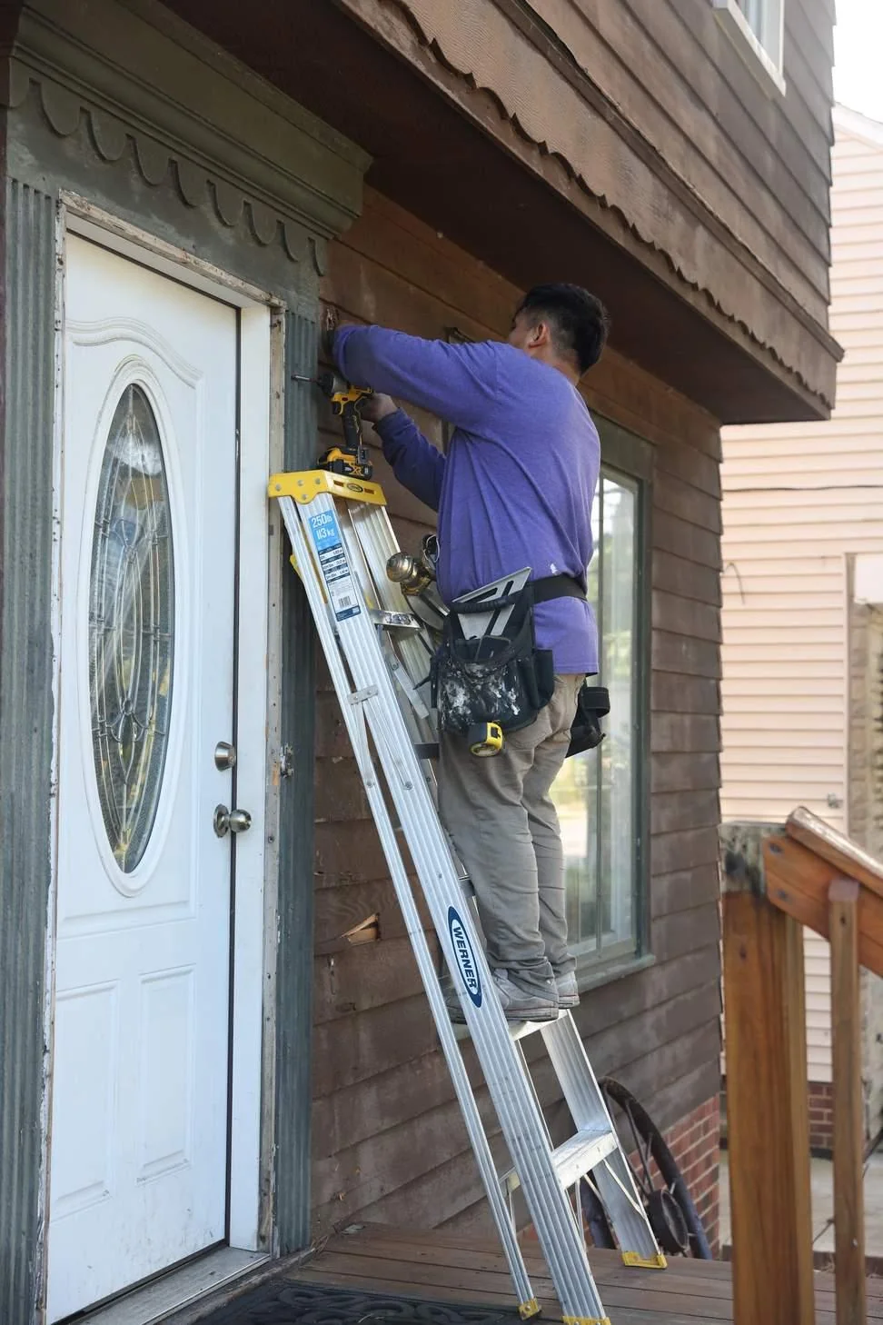 An installer on a ladder works the frame around a new white entry door, the labor most of the price pays for.