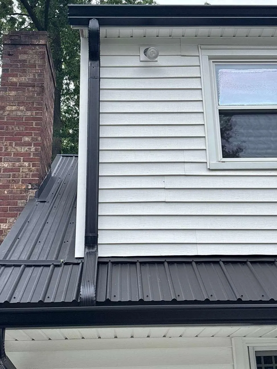 A close-up shows a dark metal roof panel meeting siding and a brick chimney, the flashing details that affect how long a roof lasts.