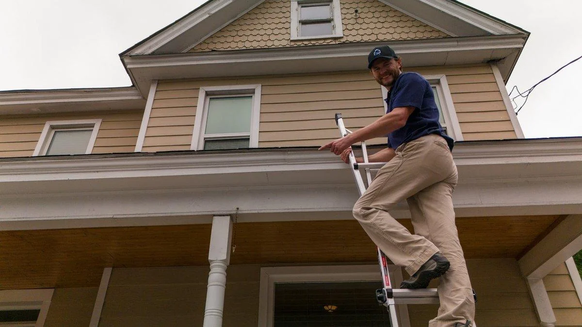 An inspector climbs a ladder to the porch roof of a tall two-story house, the kind of access that adds time to a visit.