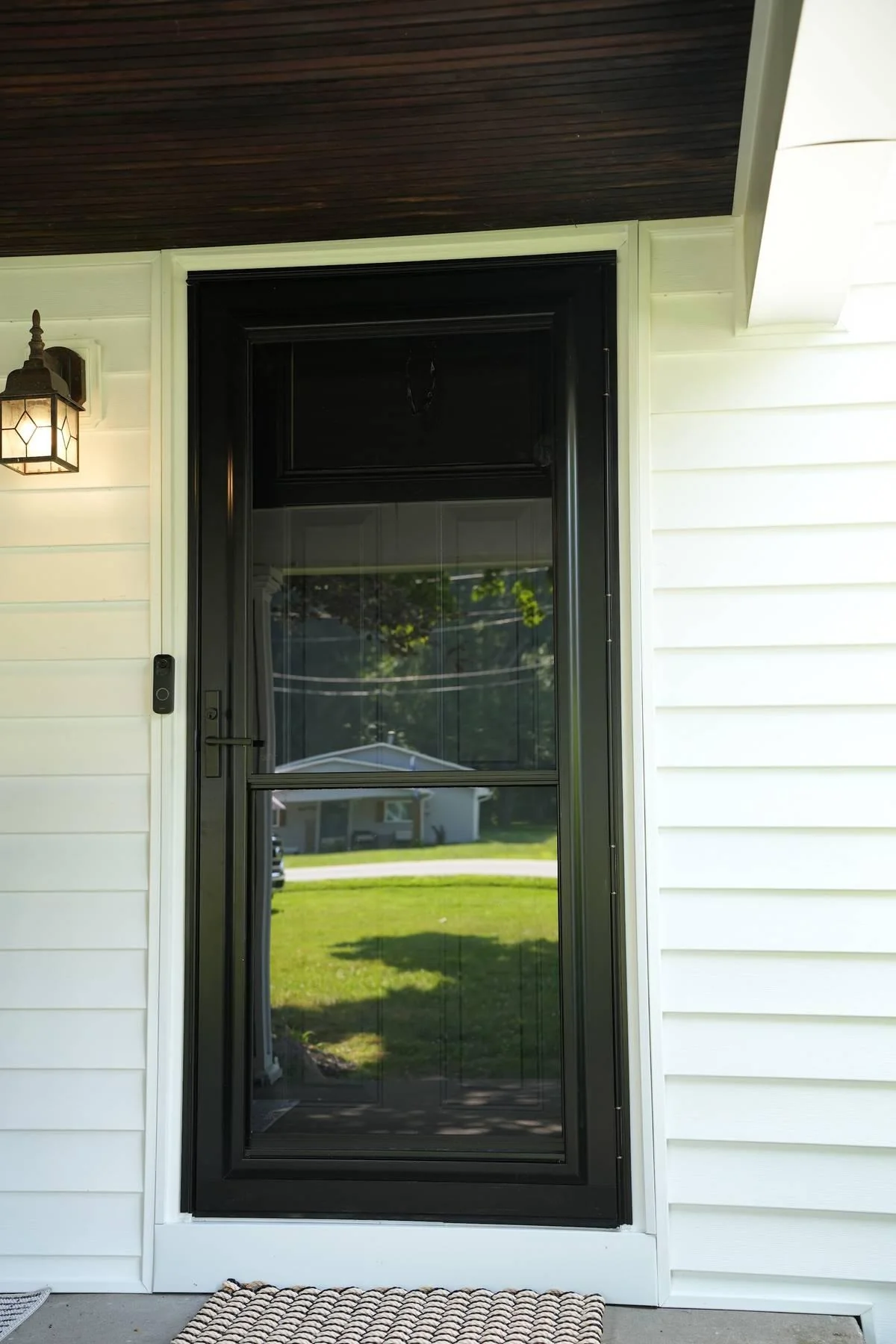 A full-view black storm door covers the entry door on a covered porch, glass panel in front and handle at the side.