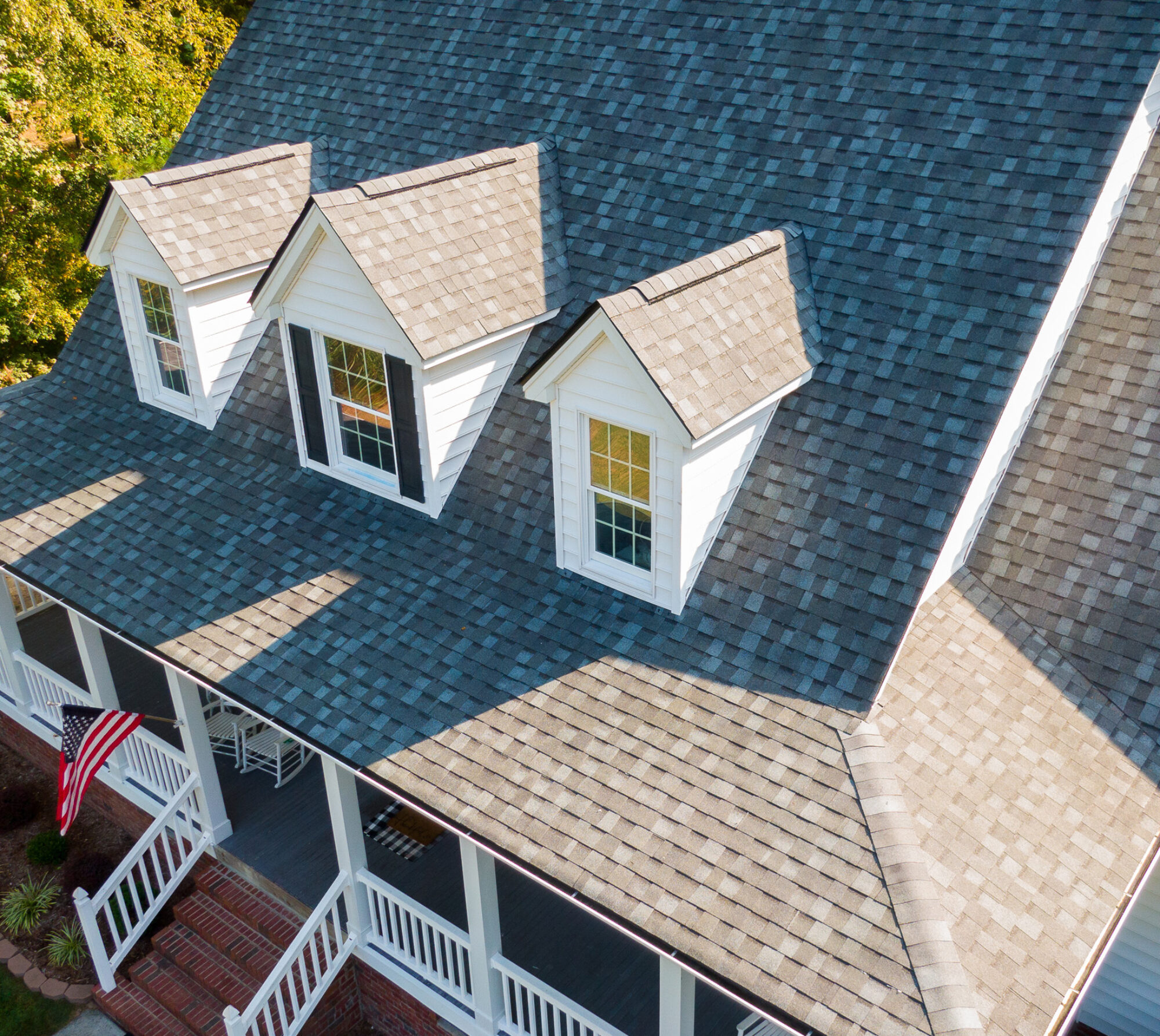 An aerial view of a suburban house with a large, sloped, shingled roof featuring dormer windows. The house has a front porch with white railings and an American flag hanging near the entrance. The property is surrounded by greenery, including tall trees in the background.