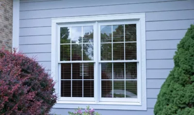 A white vinyl double-hung window with a colonial grille pattern and visible check rail set into gray siding.