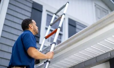 A roof inspector climbs a ladder to check the gutter and roof edge before writing up a report.